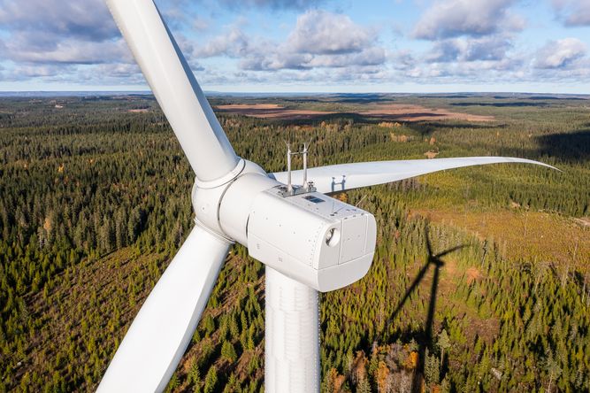 Close-up aerial view of a white wind turbine with three blades set against a backdrop of a forested landscape and partly cloudy sky.